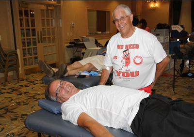 Marc A. Satalof stands beside a man laying on a blue cot, at a blood drive he chaired.